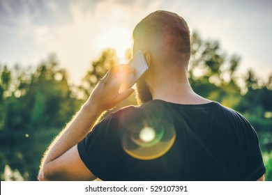 Summer Evening. Sunset. Back View. Backlight. A Young Bearded Man In A Black T-shirt Standing In A Park And Talking On A Cell Phone. A Man Uses A Digital Gadget.