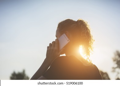 Summer Evening. Sunset. Backlight, Rear View, Young Woman, Dressed In A Black T-shirt Talking On A Cell Phone.In Background Blue Sky.Girl Holding Mobile Phone,using Digital Gadget.