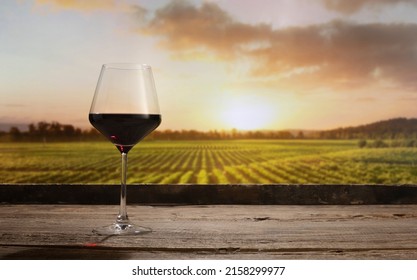 Summer Evening Time. Still Life With Red Wine Glass On Wooden Table Over Background Of Panoramic View Of Lush Vineyards At Sunset. Tasting, Festivals, Sophisticated Lifestyle And Winemaking Concept