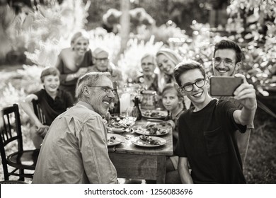 In The Summer, A Family Of Three Generations Gathered Around A Table In The Garden Sharing A Meal. A Teenager Does A Selfie With All The Guests. Black And White