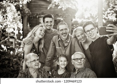 In The Summer, A Family Of Three Generations Having Fun Around A Table In The Garden Sharing A Meal. A Teenager Child Does A Selfie With All The Guests. Black And White