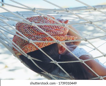 Summer Family Vacation Concept. Sideview Portrait Of Little Toddler Girl In White Dress Puts Orange Straw Cover Her Face On A Hammock In A Sandy Beach