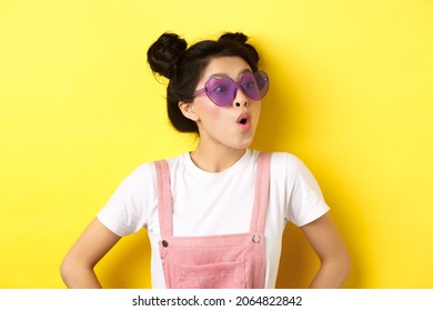 Summer And Fashion Concept. Close-up Of Surprised Asian Teen Girl In Heart-shaped Sunglasses Looking Right Side With Excited Face, Standing On Yellow Background