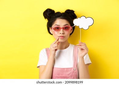 Summer And Fashion Concept. Stylish Asian Girl In Sunglasses Holding Comment Cloud On Stick And Looking Thoughtful, Thinking Or Saying Something, Standing On Yellow Background