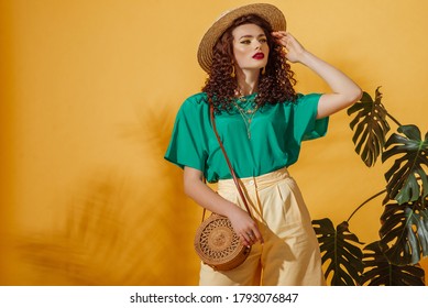 Summer fashion concept: woman wearing trendy green t-shirt, yellow bermudas shorts, straw hat, with round shoulder wicker bag, posing on yellow background. Copy, empty space for text