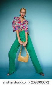 Summer fashion: confident blonde woman wearing colorful blouse, green high waist wide leg jeans, holding trendy yellow leather bag, posing on blue background. Full-length portrait