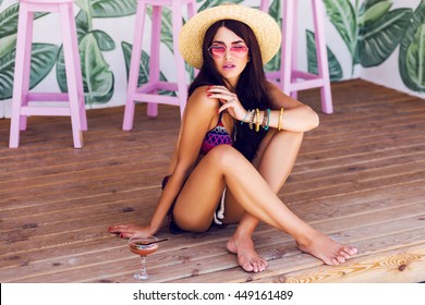 Summer fashion image of  young  woman in sexy beach bikini with tasty cocktail  sitting on wood floor in stylish  cafe .  Wearing boho accessories, straw hat, pink sunglasses. 