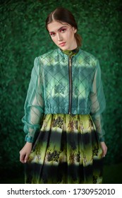 Summer Fashion. Portrait Of A Beautiful Female Model Posing In A Light Jacket And Dress With Floral Patterns On A Green Boxwood Background.