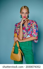 Summer fashion portrait of confident blonde woman wearing colorful blouse, green earrings, high waist jeans, holding trendy yellow leather bag, posing on blue background - Shutterstock ID 2030074352