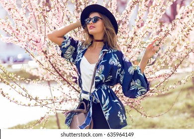 Summer fashion portrait of pretty   blonde woman posing  on amazing blooming tree background .Wearing sunglasses, casual hat , trendy blue silk kimono and  velvet choker.  Stylish  handbag.  