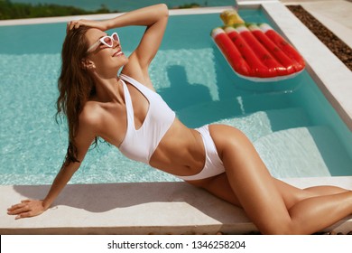 Summer fashion. Woman in swimsuit and sunglasses sunbathing near swimming pool with watermelon float. Girl model in swimwear enjoying vacation near infinity pool 