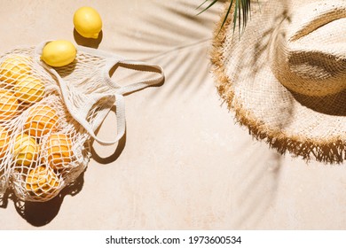 Summer flat lay on beige background. Straw hat and lemon fruits in eco friendly mesh shopping bag. Trendy palm shadow and sunlight, sun. Minimal summer travel fashion concept. - Shutterstock ID 1973600534