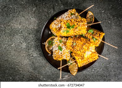Summer food. Ideas for barbecue and grill parties. Grilled corn grilled on fire. With a sprinkle of cheese (elotes), hot chili pepper, lemon. On a dark stone table, black plate Copy space top view