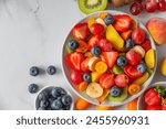 Summer fresh fruit salad in a bowl on white marble background. Top view with copy space. Healthy food for breakfast. Mixed strawberries, grapes, banana, kiwi, blueberries, peach, citrus for diet lunch