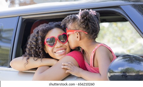 Summer Fun! Happy Girls Or Sisters With Heart Shaped Sunglasses In Car Window. Younger Girl Kisses Older Girl On Cheek In Celebration Of Love, Summer Holiday, Road Trip Or Valentines Day. Copy Space.