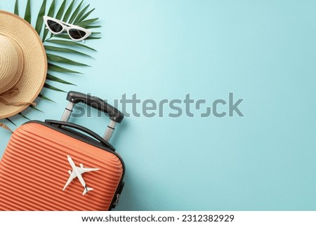 Summer getaway concept. Top view of an orange suitcase, small airplane figurine, beach essentials, sunglasses, sunhat, palm leaves on pastel blue background, providing space for text or promo content
