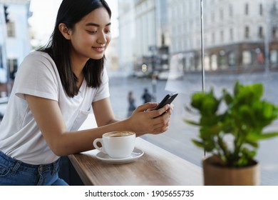 Summer girl sitting in cafe with cup of coffee, messaging on smartphone. Young woman using mobile phone in restaurant while drinking cappuccino near window.