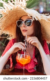 Summer Girl In Sunglasses, Straw Hat And Red Polka-dot Dress Sitting On The Terrace With Aperol Spritz Drink. Young Woman Portrait