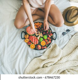 Summer Healthy Raw Vegan Clean Eating Breakfast In Bed Concept. Young Girl Wearing Pastel Colored Home Clothes Taking Cherries From Tray Full Of Fresh Seasonal Fruit. Top View, Square Crop, Copy Space