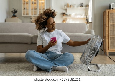 Summer heat at home. Happy curly African American woman sitting with smartphone on floor enjoying cool fresh air blowing from electric fan, chatting on mobile phone relaxing in front of air cooler - Powered by Shutterstock