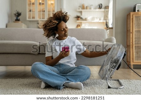 Summer heat at home. Happy curly African American woman sitting with smartphone on floor enjoying cool fresh air blowing from electric fan, chatting on mobile phone relaxing in front of air cooler