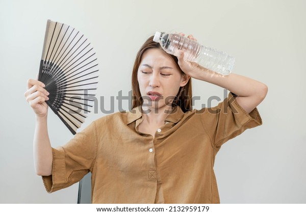 Summer heat stroke, hot weather, tired asian young\
woman sweaty and thirsty, refreshing with hand in blowing, wave fan\
to ventilation, holding cold water bottle tap her body when\
temperature high.