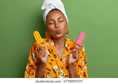 Summer Is Here. Lovely Pleased Woman Stands With Closed Eyes, Folded Lips, Holds Two Delicious Ice Creams, Wears Yellow Dressing Gown With Flower Print And Towel Wrapped On Head, Isolated On Green