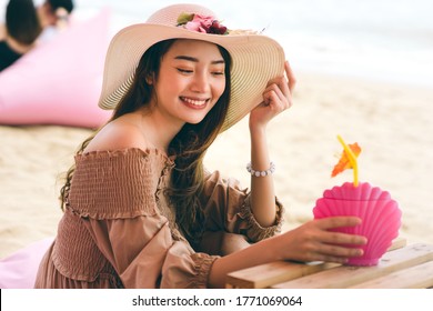 Summer holiday concept. Young adult asian woman relax at the beach sitting on pink cafe chair. Her face with happy smile wear hat and grab drink beverage. Outdoor at day light. - Powered by Shutterstock