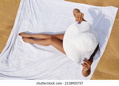 Summer holiday fashion concept. A tanned woman wears a sun hat on the beach on a white blanket on the sand.