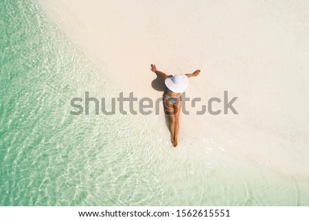 Summer holiday fashion concept - tanning girl wearing sun hat at the beach on a white sand shot from above.Top view from drone. Aerial view of slim woman sunbathing lying on a beach in Maldives.