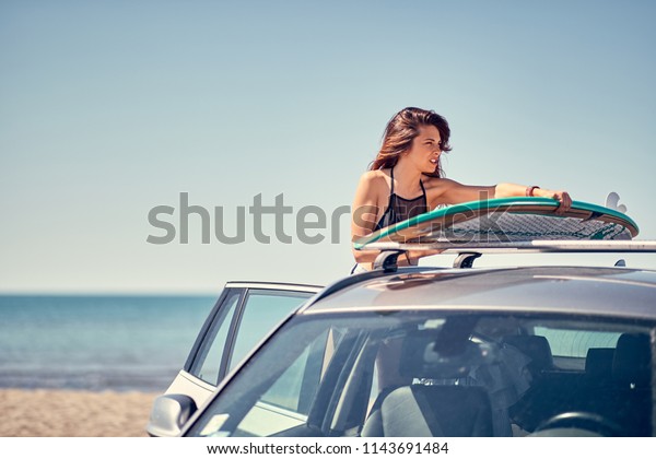 Summer holiday road trip vacation- Young smiling\
surfer girl at the beach with her surfboard on car Vacation.\
Extreme Sport\
