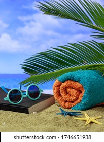 Summer holidays concept. Sunglasses, book and towel on a sandy beach with palm tree, sun, clouds on blue sky, blur sea background. Vertical photo, closeup view.