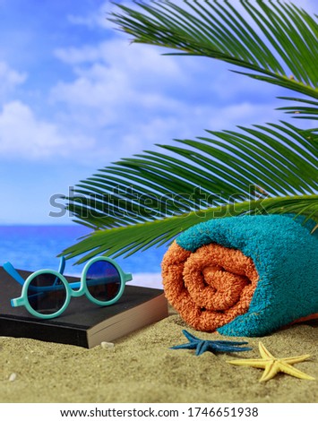 Summer holidays concept. Sunglasses, book and towel on a sandy beach with palm tree, sun, clouds on blue sky, blur sea background. Vertical photo, closeup view.