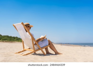 Summer holidays on the Baltic Sea. Woman of European beauty in white tunic and beige hat reading book sitting on deckchair on beach with white sand on beautiful sunny day. Relaxing on beach. Side view - Powered by Shutterstock