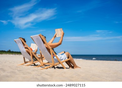 Summer holidays on beach with book. Two women of European beauty dressed in bikini and shorts reading books sitting on deckchairs on beach with white sand on beautiful sunny day. Relaxing on beach. - Powered by Shutterstock