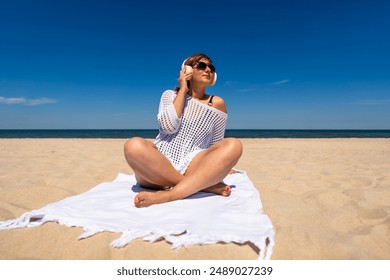 Summer holidays on beach with music. Beautiful young woman in black swimsuit and white tunic listening to music in headphones and sitting cross-legged on towel on white sandy beach. Front view - Powered by Shutterstock