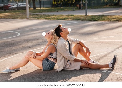 Summer Holidays And People Concept - Happy Young Couple Sitting Back To Back And Blowing Bubble Gums On Basketball Playground