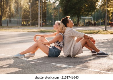 Summer Holidays And People Concept - Happy Young Couple Sitting Back To Back On Basketball Playground