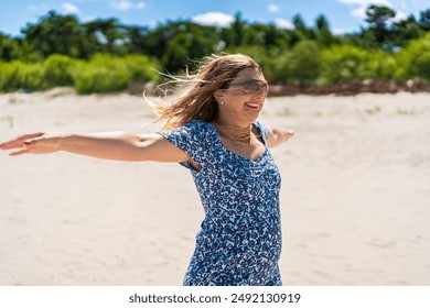 Summer holidays. Portrait of middle-aged laughing woman with long hair in blue dress and sunglasses with outstretched arms on sandy beach on sunny day. Energy, happiness, joy of life and youth - Powered by Shutterstock