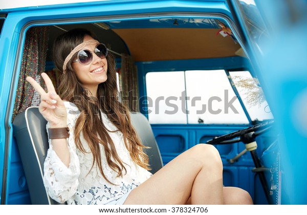 summer holidays, road trip, vacation, travel and\
people concept - smiling young hippie woman showing peace gesture\
in minivan car