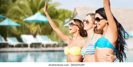 summer holidays, travel, people and vacation concept - happy young women in bikinis and shades hugging and waving hands over swimming pool background