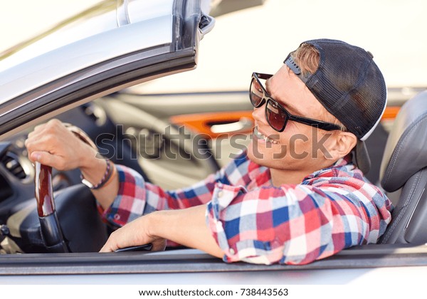 summer holidays, travel, road trip and people concept
- happy smiling young man in sunglasses and cap driving convertible
car