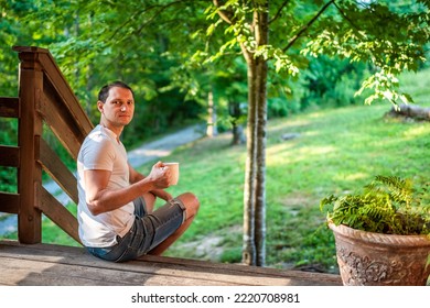 Summer house porch with man sitting on steps of house in front or back yard morning wooden cabin cottage drinking coffee or tea from mug looking back - Powered by Shutterstock