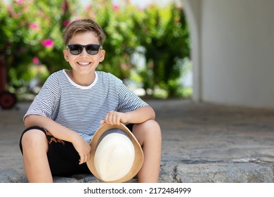 Summer Kid. Portrait Of Handsome Schoolboy Wearing Sunglasses Sitting Outdoor With Straw Hat In His Hands At Sunny Day With Nature Background.