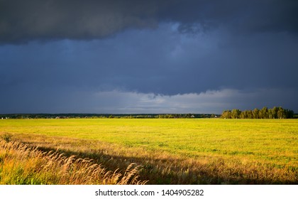 Summer Landscape With A Dark Pre-storm Sky And A Bright Yellow Field, Divided Into Two Parts By A Thin Line Of Forest On The Horizon With The Roofs Of Village Houses On Its Background