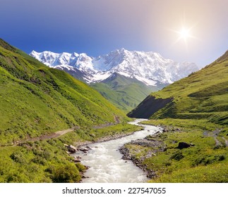 Summer Landscape With River And Mountain Snow. Peak Shkhara Zemo Svaneti, Georgia. The Main Caucasian Ridge