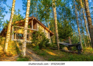 Summer Landscape With Wooden Eco Cottage And Hammock In Forest In Evening At Sunset. Facade Of Log Cabin Among Birch Trees. Staycation In Fresh Air, Beautiful Location, Rest In Country House.