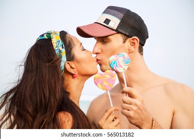 Summer lifestyle close up portrait of happy kissing couple in love. Holding in their hands lollipops in shape of heart. On honeymoon. Interracial couple, Asian woman, Caucasian man. Bright colors 