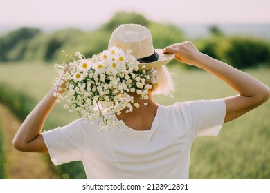 Summer Lifestyle Concept. Trendy adult woman in stylish summer dress feeling free in the field with wild flowers in sunshine. Model in sraw hat. Rear view.
