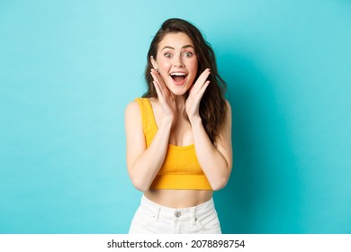 Summer And Lifestyle Concept. Young Cheerful Woman Look With Surprised Face At Camera, React To Awesome Good News, Smiling And Cheering, Standing Against Blue Background